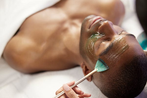 Man lying down with green gel mask applied to face using a brush in a clinical setting