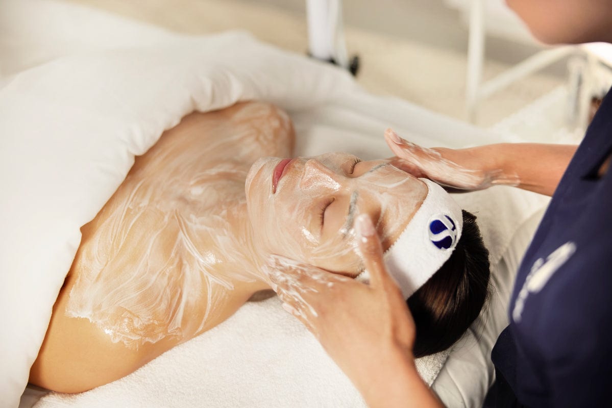 Person receiving a facial with white cream on face and shoulders, wearing an iS Clinical headband