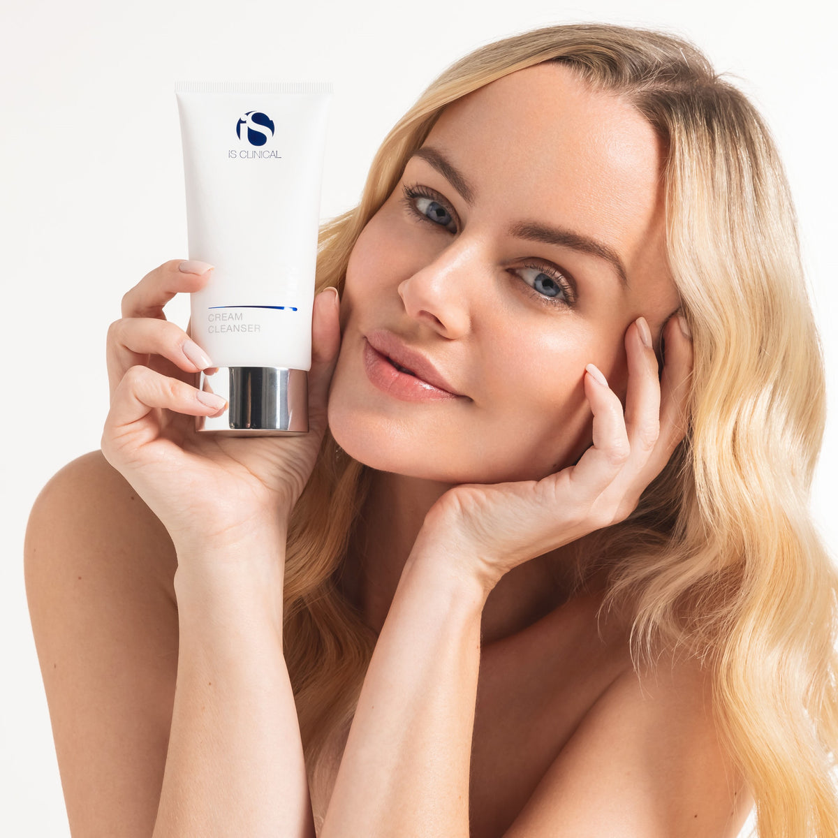 Woman with clear skin holding iS Clinical Cream Cleanser tube near her face against white background