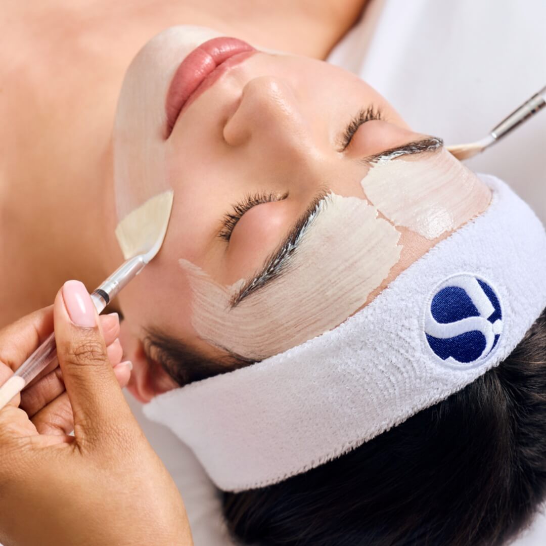 Close-up of a woman with eyes closed wearing a white headband, having a white facial mask applied with brushes