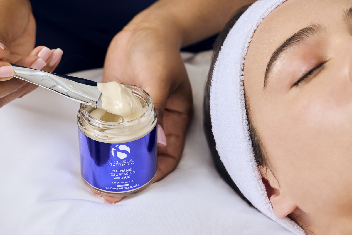 Hand holding iS Clinical Intensive Resurfacing Masque jar with spatula applying cream near closed eyes