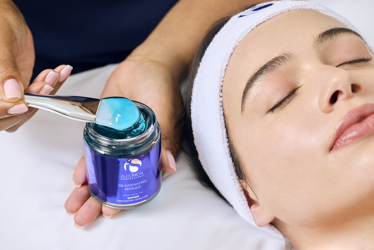 Close-up of a person with eyes closed wearing a white headband, blue iS Clinical Rejuvenating Masque being scooped with a silver spatula