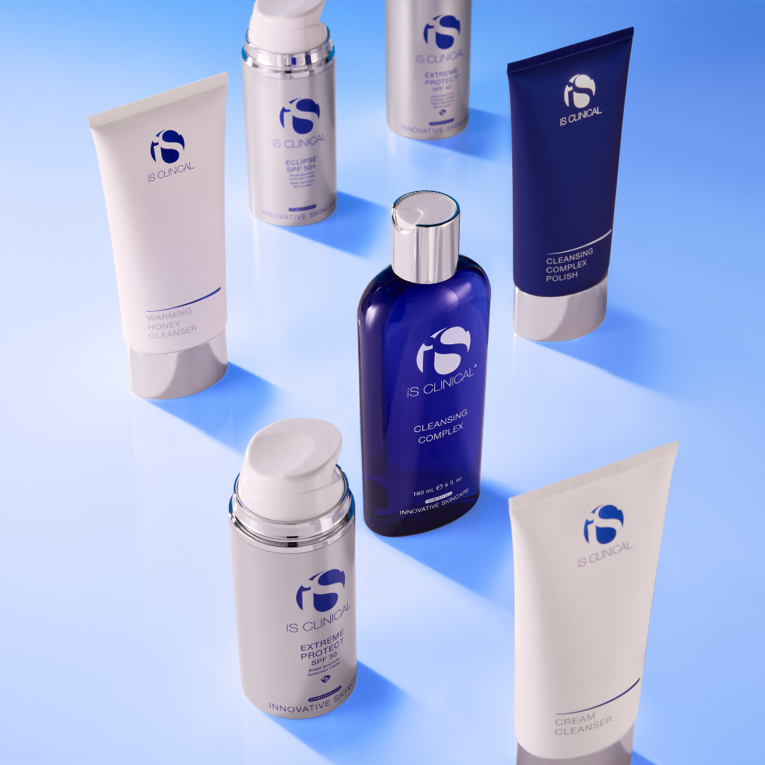 Seven iS Clinical skincare products including Warming Honey Cleanser, Cleansing Complex, Cleansing Complex Polish, Cream Cleanser, and Extreme Protect SPF 30 and 50+ bottles on blue reflective surface
