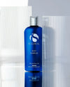 Blue bottle of iS Clinical Body Complex with silver cap against textured white and grey background