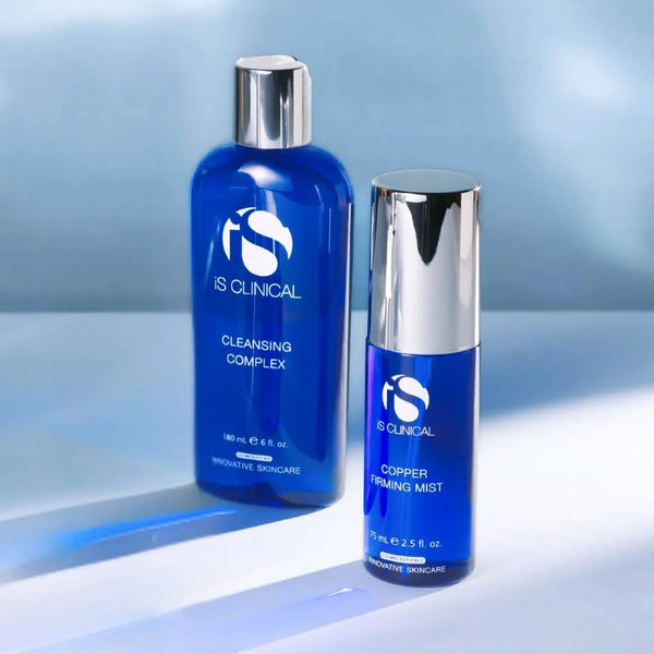 Two blue iS Clinical bottles with silver caps, Cleansing Complex and Copper Firming Mist, on light blue surface