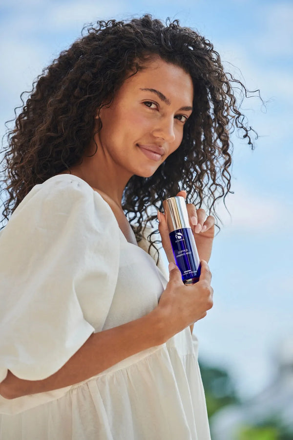 Woman holding iS Clinical Copper Firming Mist in blue bottle with silver cap against sky background