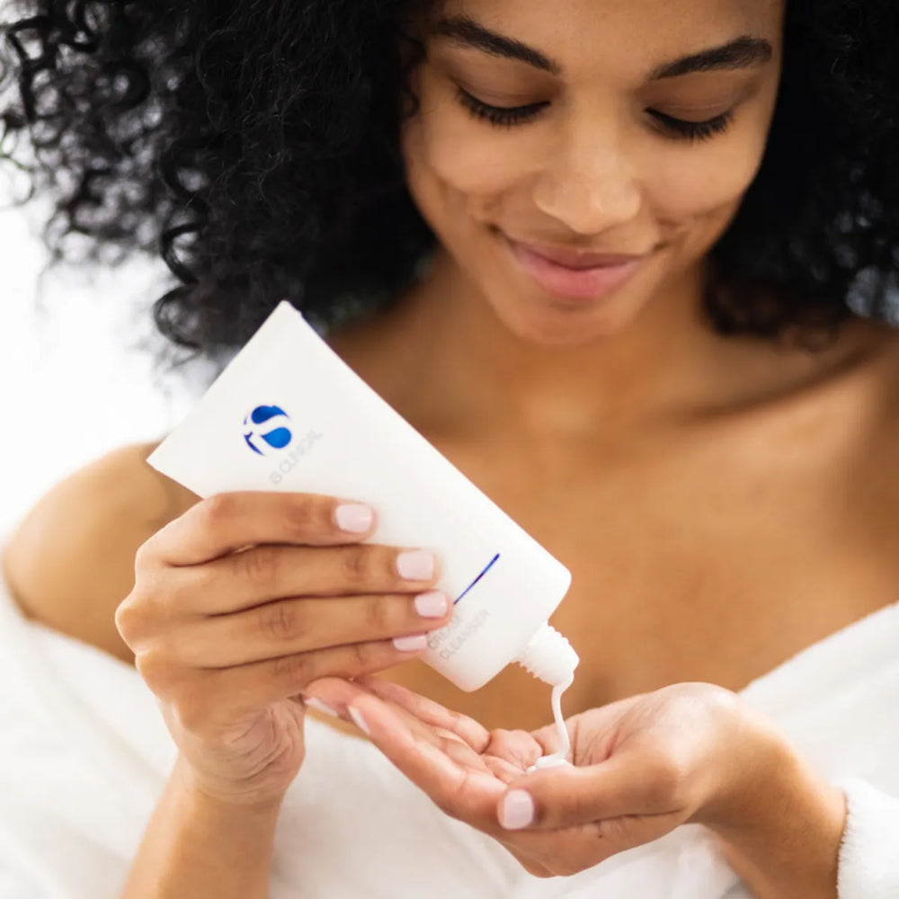 Woman dispensing white cream cleanser from white iS Clinical tube onto palm