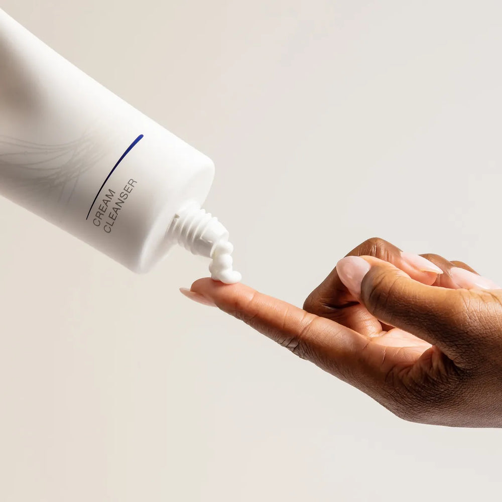 White tube of iS Clinical Cream Cleanser dispensing white cream onto fingertip against neutral background