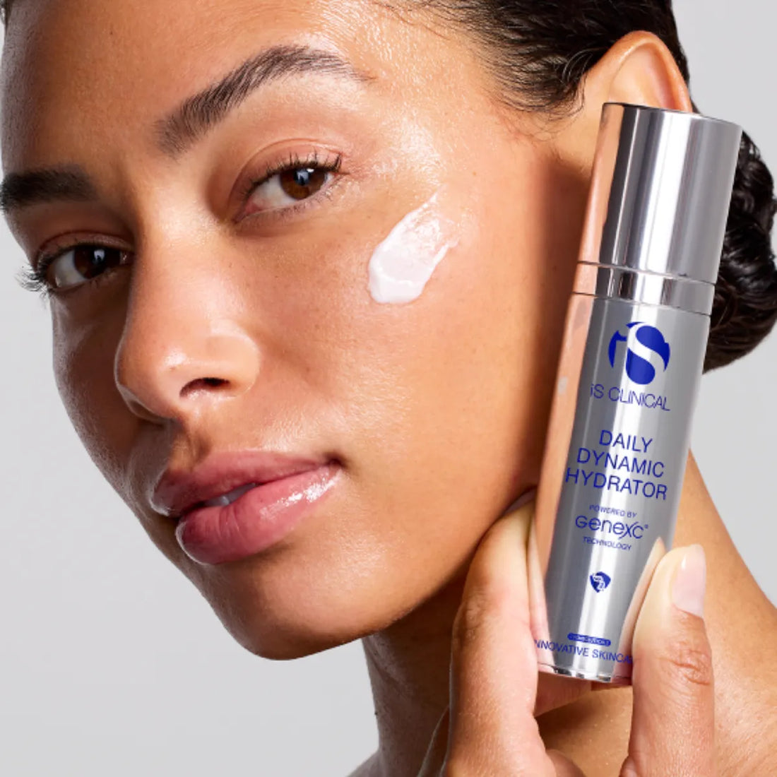 Close-up of woman with cream on cheek holding iS Clinical Daily Dynamic Hydrator silver bottle