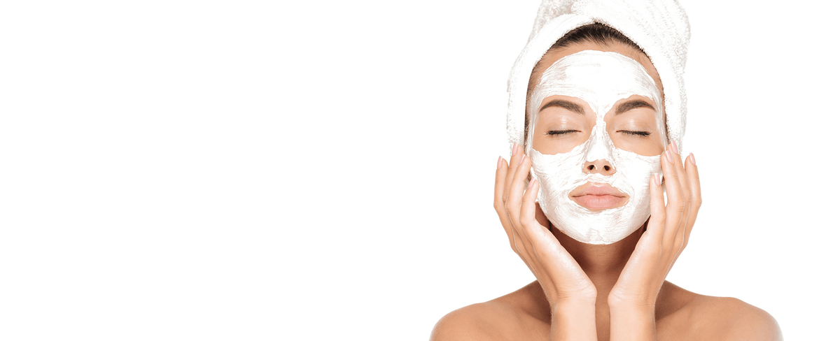 Woman with white facial mask applied, eyes closed, hands gently touching face, hair wrapped in towel