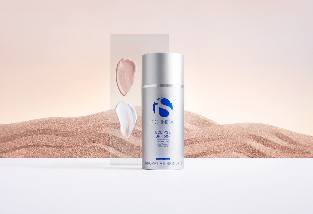 iS Clinical Eclipse SPF 50+ sunscreen bottle with silver cap and blue logo, two cream swatches on transparent panel, sandy textured background