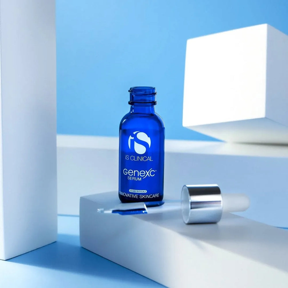 Blue glass bottle of iS Clinical GeneXC Serum with silver dropper cap on white block against blue background
