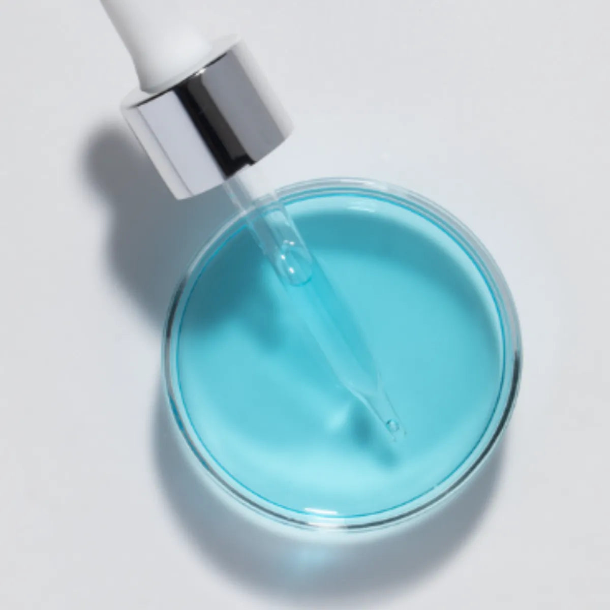 Dropper with silver collar dispensing blue serum into clear dish on white background