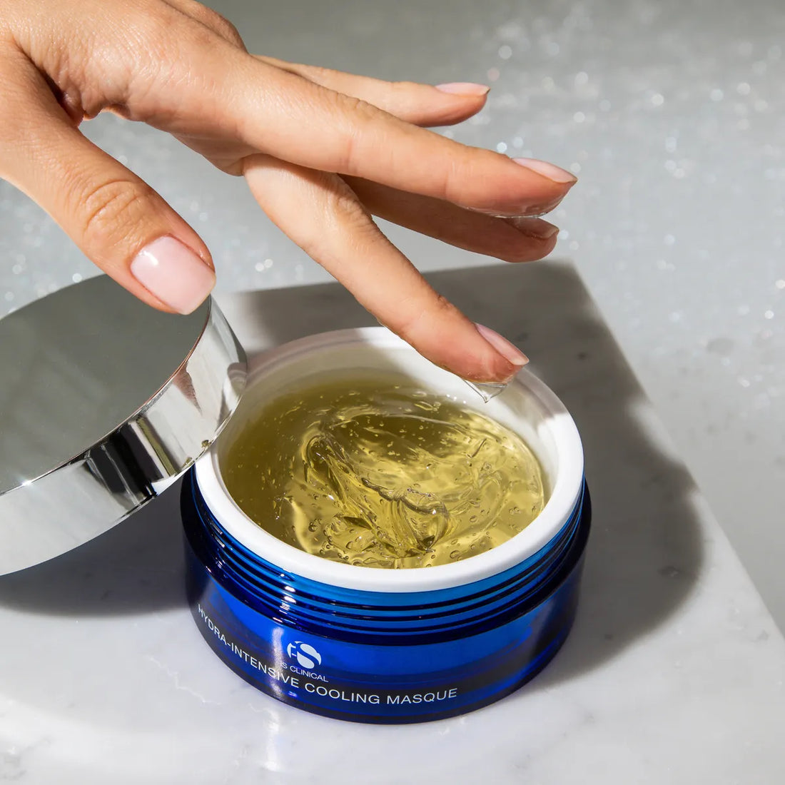Hand scooping clear gel from an open blue jar of iS Clinical Hydra-Intensive Cooling Masque