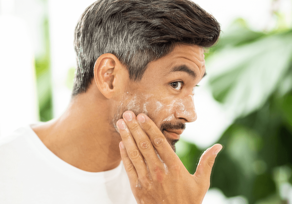 Man applying foaming cleanser to cheek with fingers, green foliage background