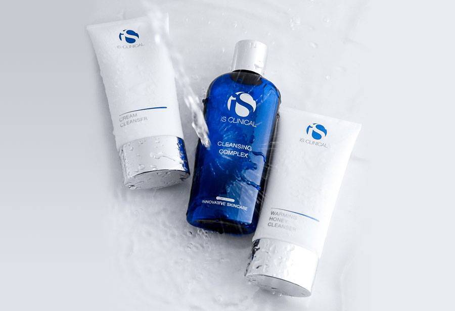 iS Clinical Cream Cleanser and Warming Honey Cleanser tubes with Cleansing Complex blue bottle under running water on white background