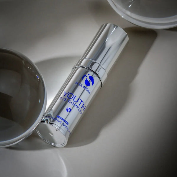 Silver cylindrical bottle of iS Clinical Youth Eye Complex with blue text on a reflective surface