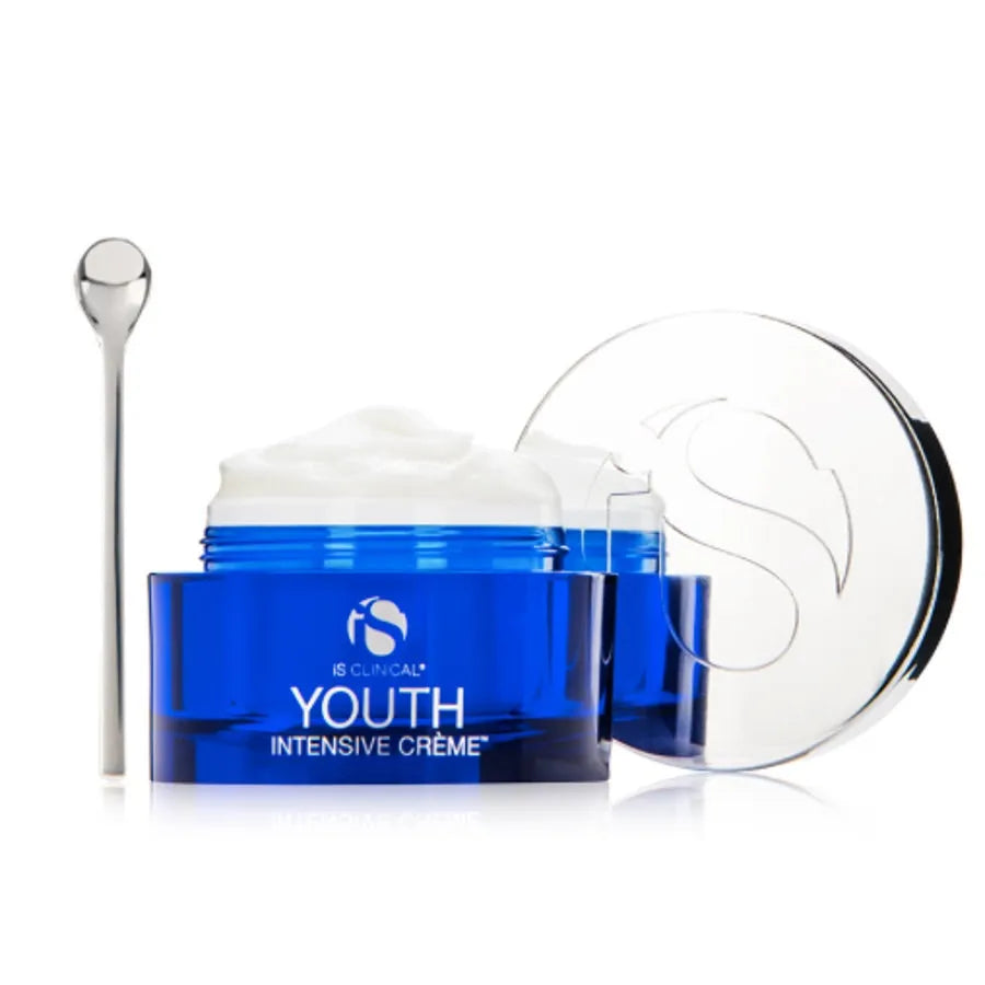 Open blue jar of iS Clinical Youth Intensive Crème with white cream and silver spatula on white background